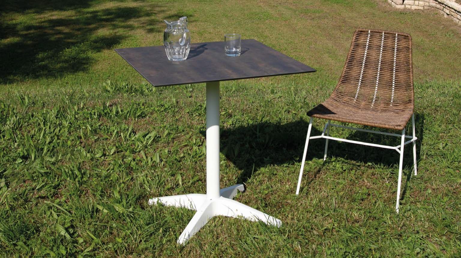 Tavolino da esterno con due bicchieri accanto a una sedia intrecciata sull'erba, al sole.