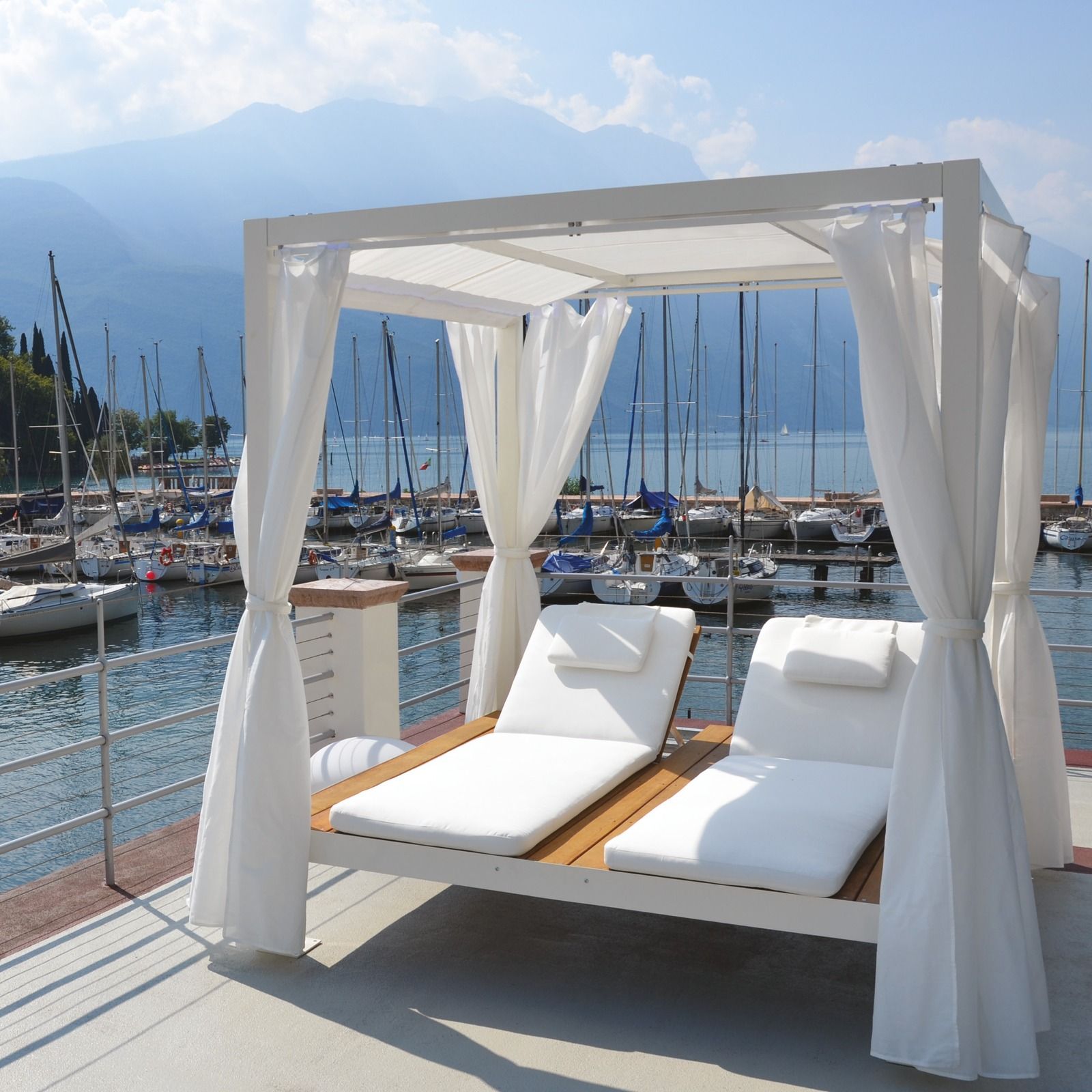 Cabina bianca con lettino prendisole su un molo del porto turistico, con vista sulle barche e sull'acqua azzurra sotto un cielo soleggiato.
