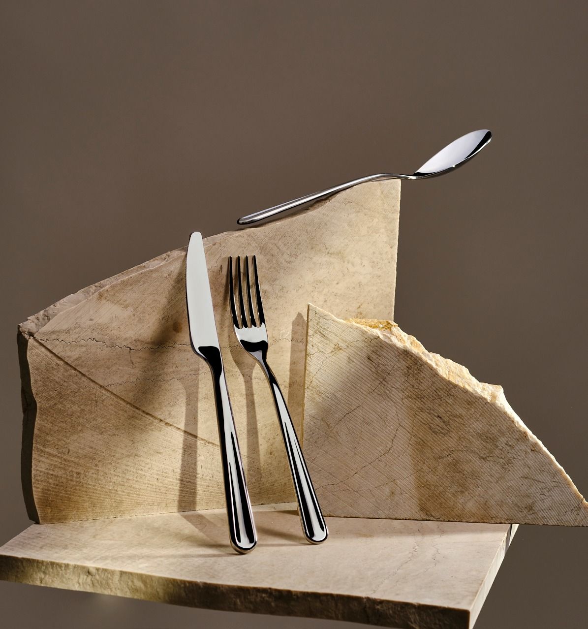 Forchetta, coltello e cucchiaio in equilibrio su un tovagliolo beige piegato, su uno sfondo marrone.