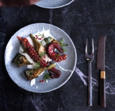 Antipasto a base di polpo servito su un piatto grigio con forchetta e coltello su un tavolo di marmo scuro.