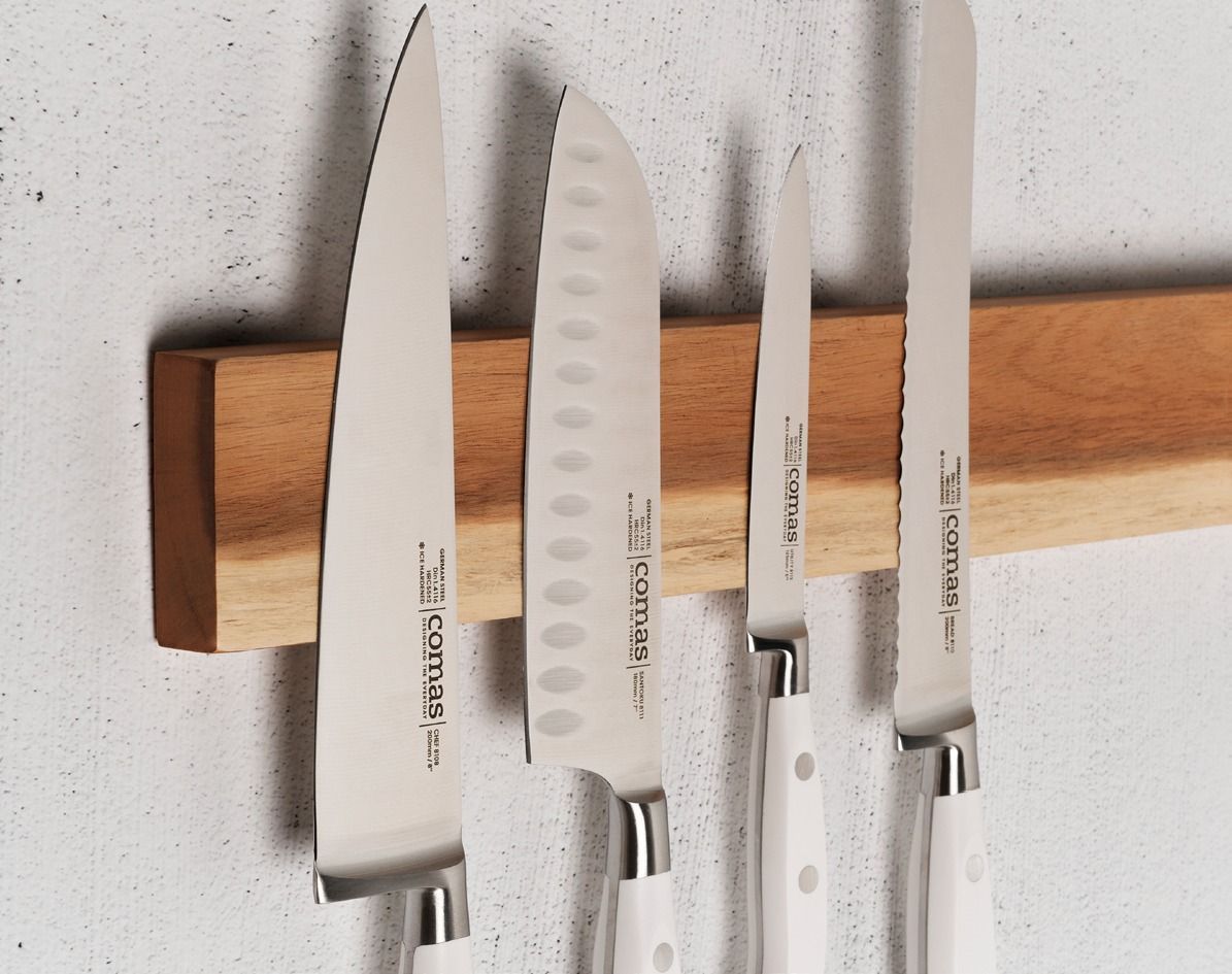Quattro coltelli da cucina argentati appesi a un supporto magnetico in legno su una parete bianca.