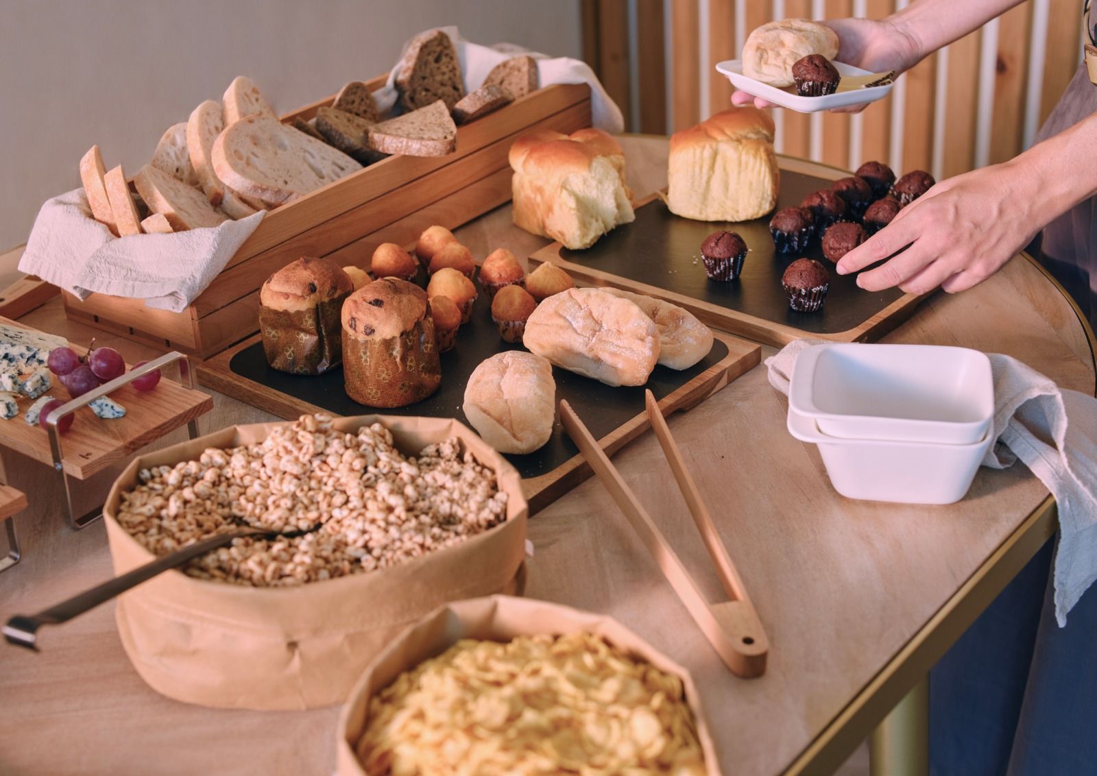 Un tavolo con un assortimento di pane, pasticcini e cucchiai, e delle mani che servono il cibo a un buffet.