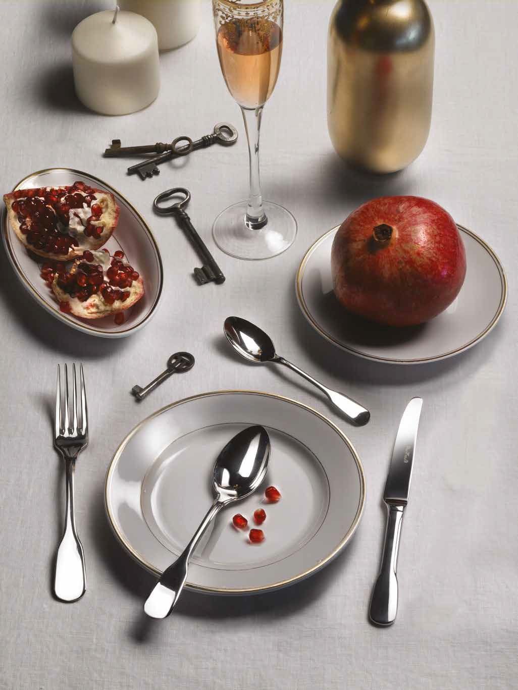 Tavola apparecchiata in modo formale con melograno, flûte da champagne e posate su piatti bianchi.