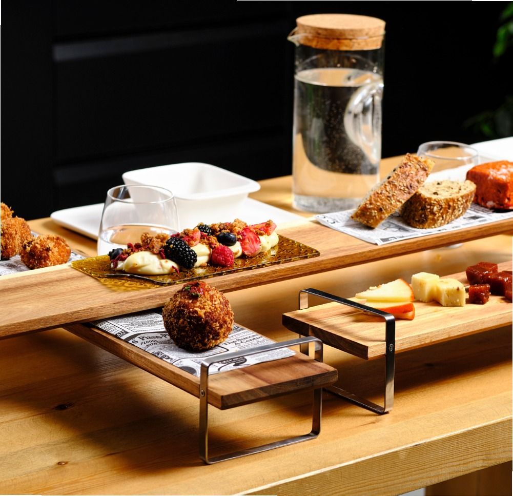 Antipasti moderni serviti su vassoi di legno con caraffa di vetro e ciotoline su un tavolo.