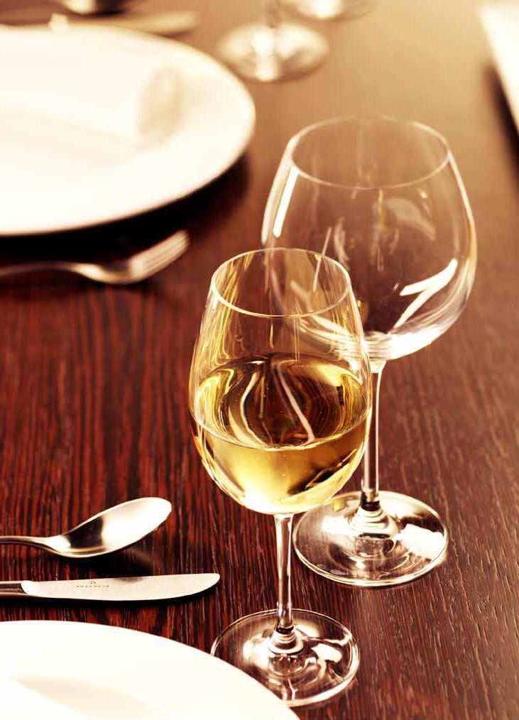 Su un tavolo di legno di un ristorante, accanto a piatti e cucchiai, ci sono due calici da vino, uno dei quali riempito di vino bianco.