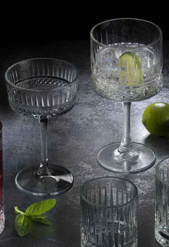 Eleganti calici in vetro trasparente e un bicchiere con ghiaccio e lime su un tavolo scuro, con foglie di menta nelle vicinanze.