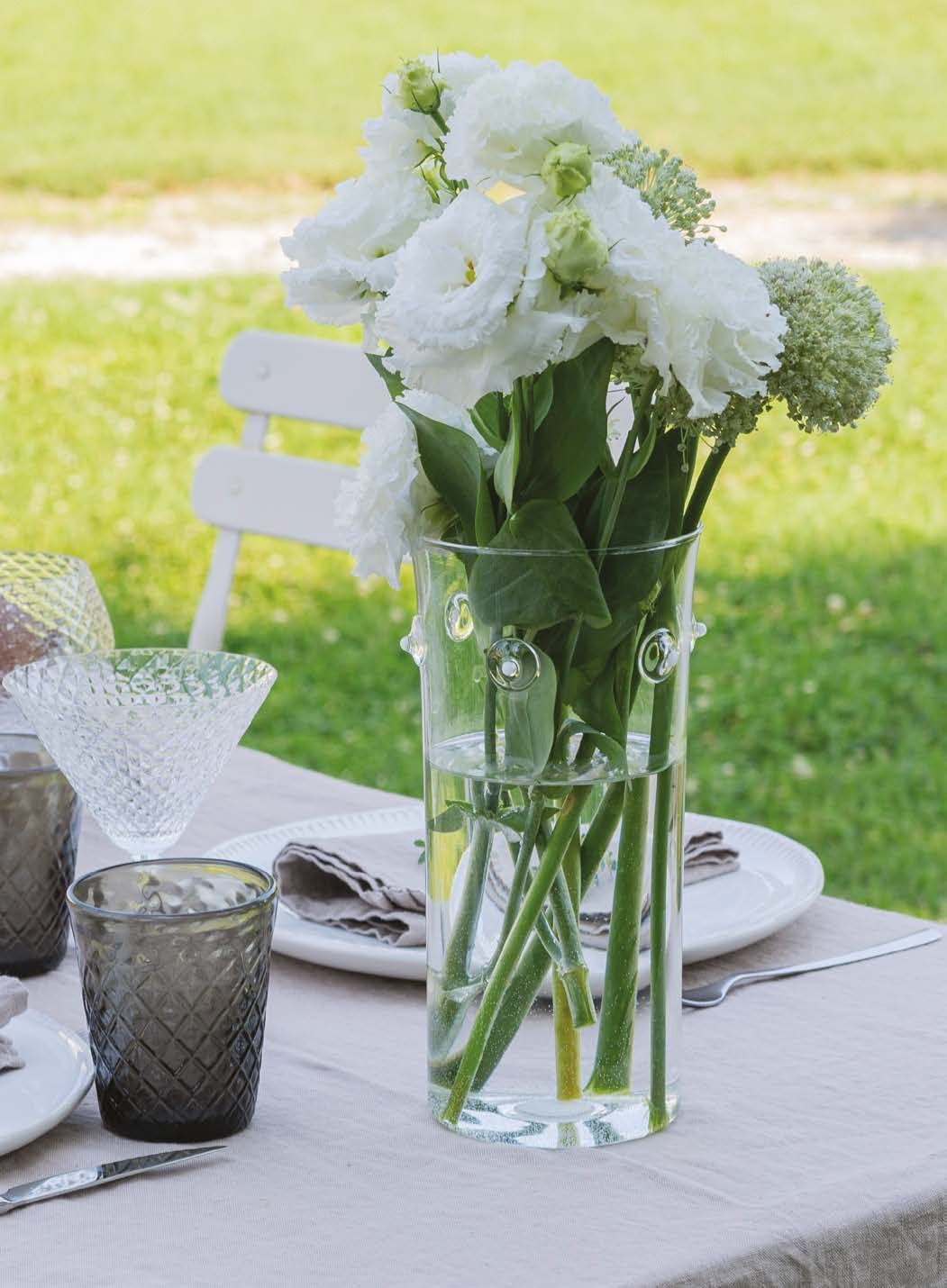 Fiori bianchi in un vaso di vetro su un tavolo all'aperto, con piatti bianchi e un prato verde sullo sfondo.