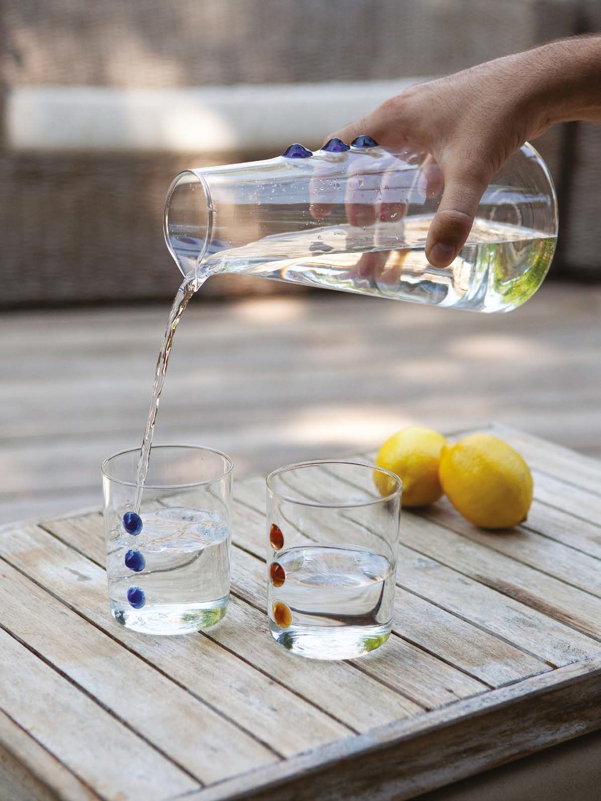 Una mano versa acqua da una brocca in un bicchiere su un tavolo di legno accanto a dei limoni.