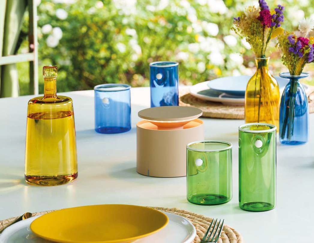 Stoviglie colorate in vetro e ceramica disposte su un luminoso tavolo da pranzo all'aperto.