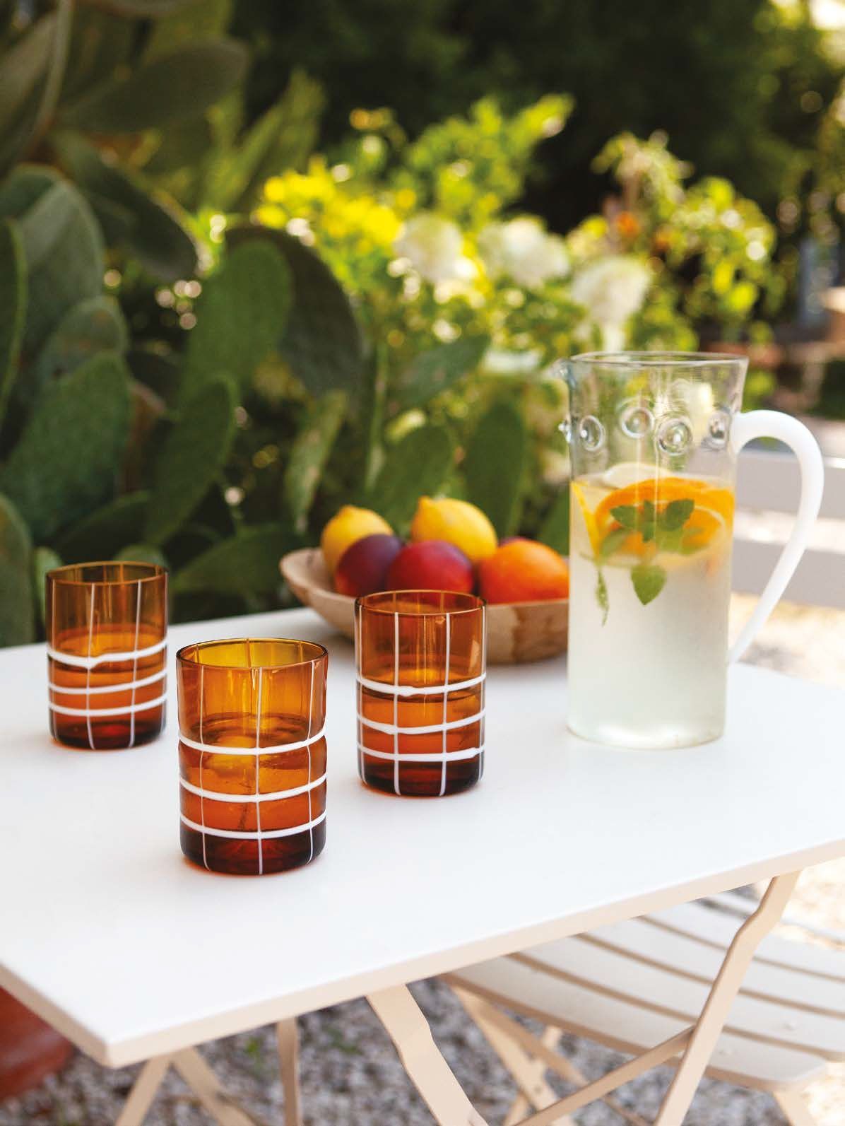 Tavolo all'aperto con bicchieri a righe, caraffa per bevande agli agrumi e una ciotola di frutta in un giardino soleggiato.