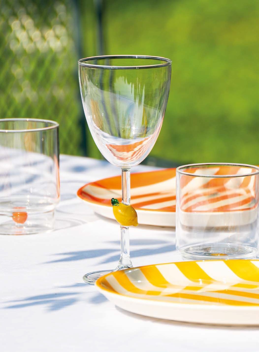 Tavolo all'aperto illuminato dal sole con piatti a righe, bicchieri trasparenti e un calice di vino vuoto su una tovaglia bianca.