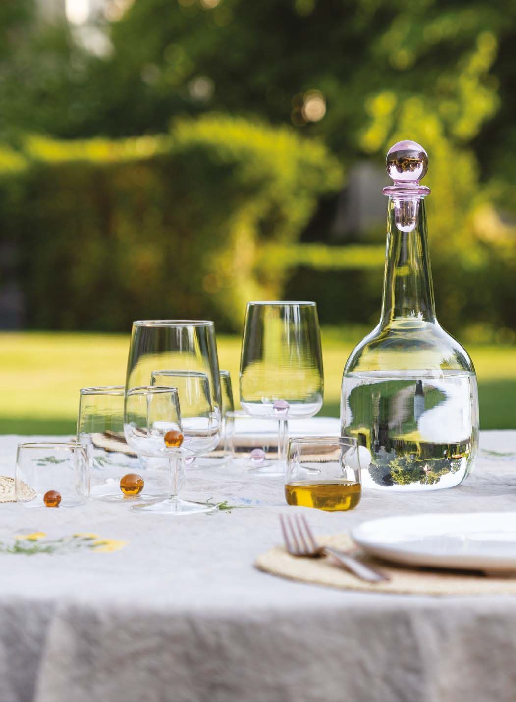 Tavolo da esterno apparecchiato con bicchieri da vino, caraffa e coppette da dessert su una tovaglia bianca.
