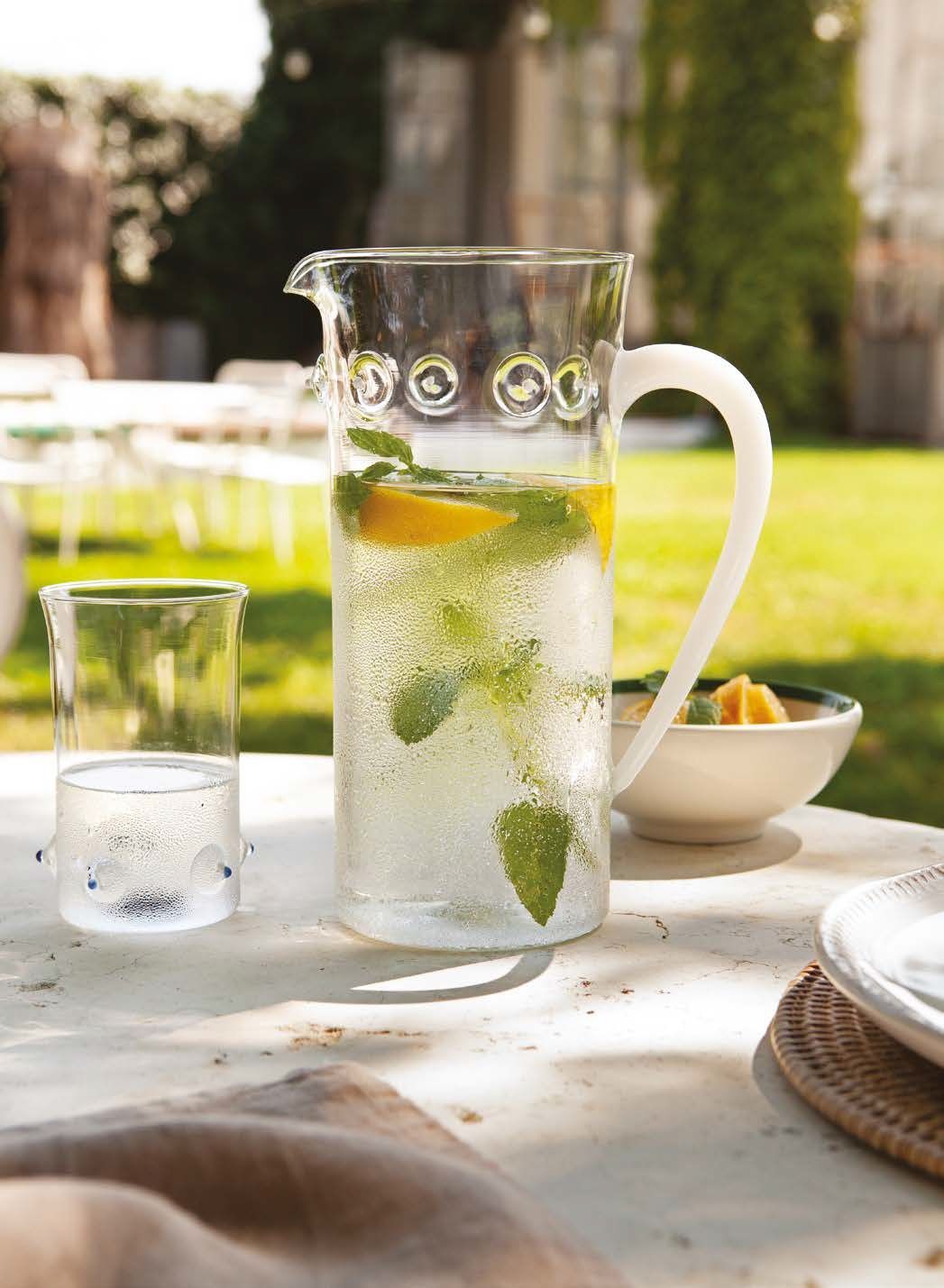 Una brocca di acqua aromatizzata al limone e menta su un tavolo all'aperto, accanto a un bicchiere e una ciotola, in un giardino soleggiato.