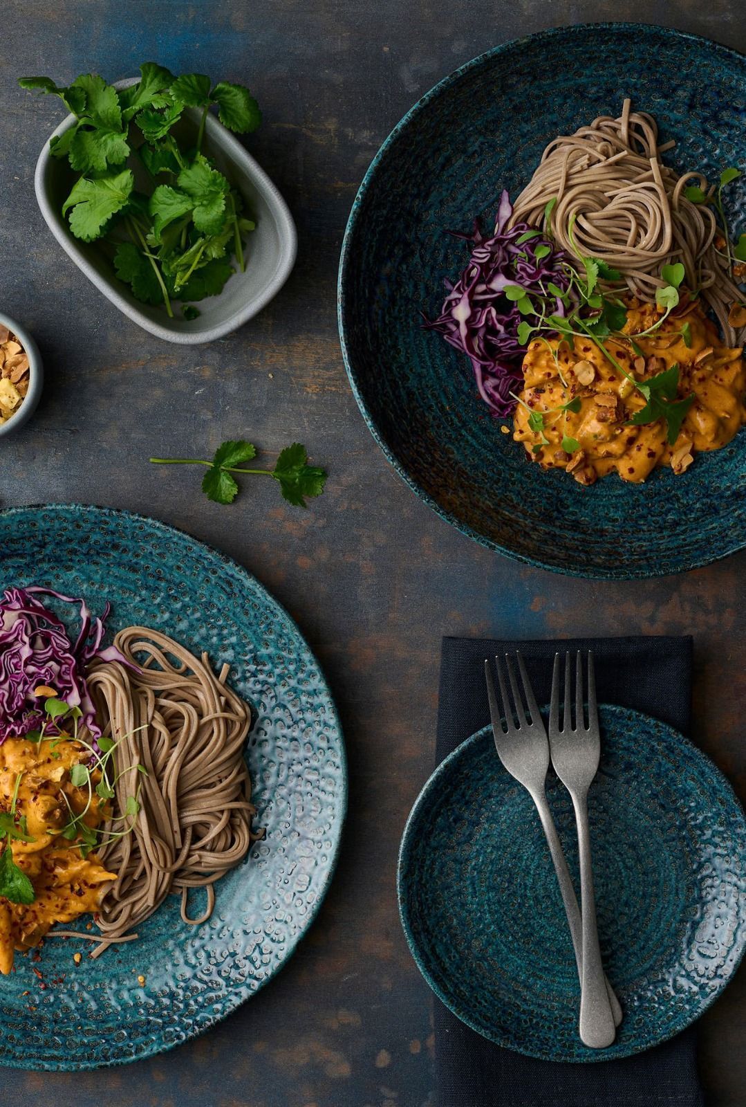 Vista dall'alto di due ciotole di noodles con verdure e cavolo viola su un tavolo scuro, con una piccola ciotola di erbe aromatiche e piattini.