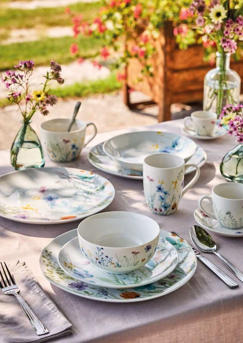 Tazze e piattini con motivi floreali disposti su un tavolo all'aperto, con fiori sullo sfondo.