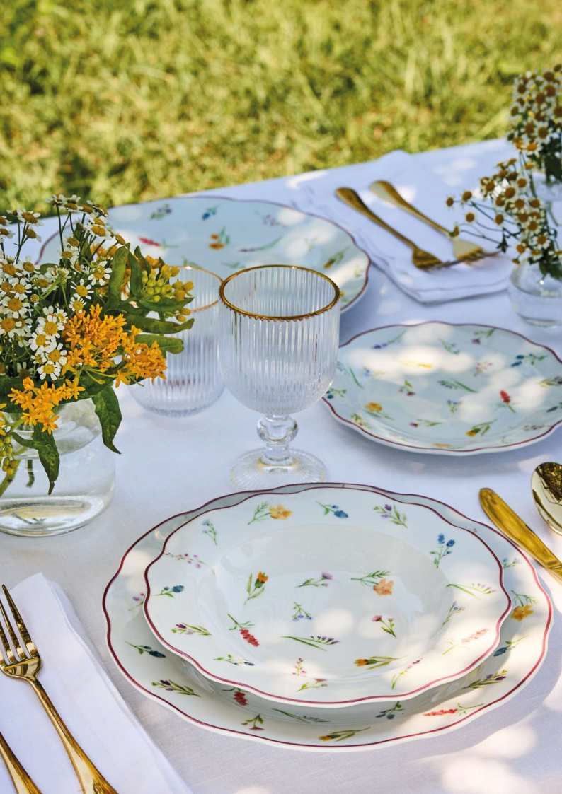 Elegante allestimento da tavola per esterni con piatti floreali, posate dorate, bicchieri di cristallo e fiori gialli su una tovaglia bianca.
