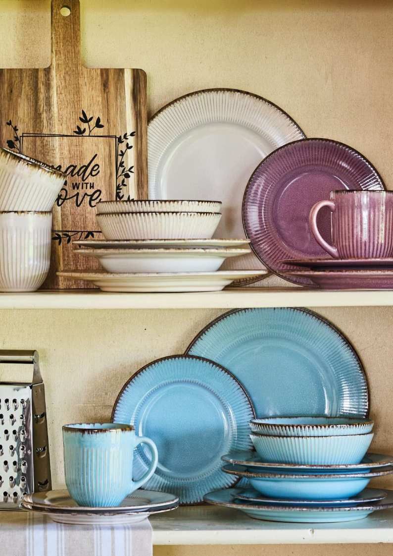 Stoviglie dai colori pastello disposte sugli scaffali della cucina, con piatti e tazze blu, viola e bianchi.