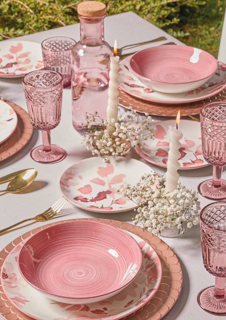 Allestimento della tavola con fiori rosa, stoviglie, bicchieri, candele e fiori bianchi all'aperto.