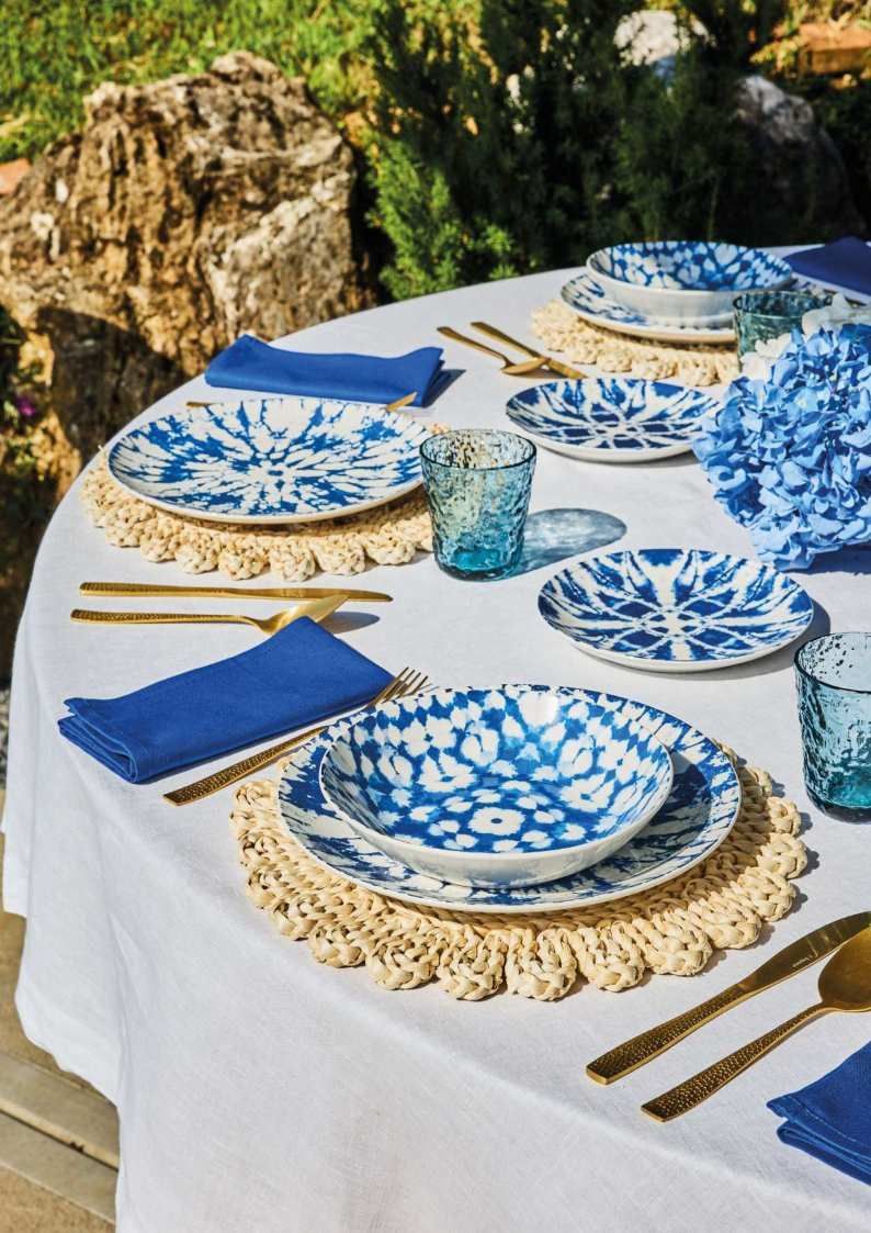Servizio da tavola per esterni blu e bianco con piatti, tovaglioli e bicchieri su una tovaglia bianca.