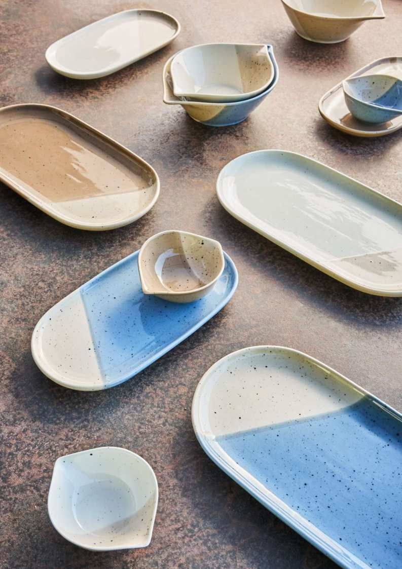 Piatti e ciotole in ceramica assortiti nei colori crema, blu e con finitura maculata, disposti su un tavolo.