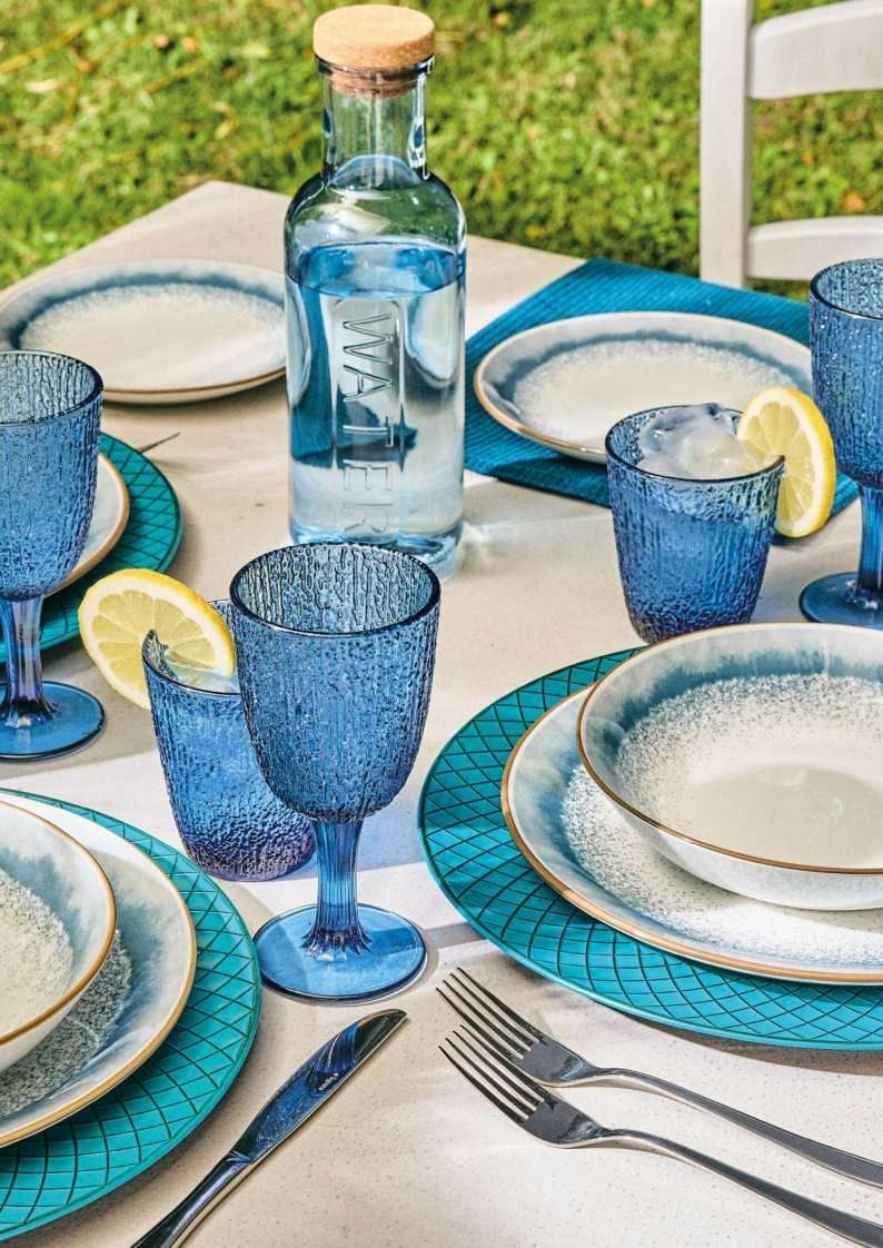 Servizio da tavola in vetro blu con piatti, calici, fette di limone e una bottiglia d'acqua su un tavolo da patio.
