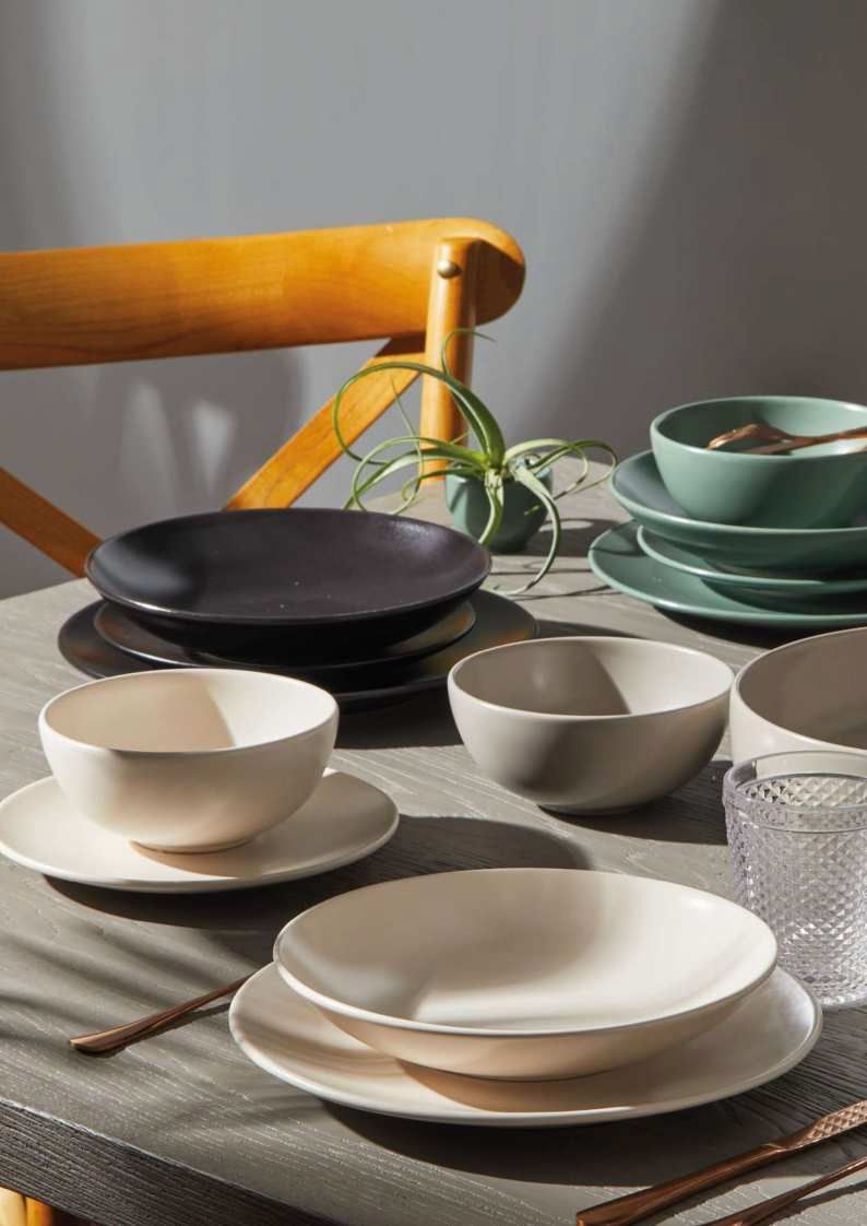 Servizio da tavola in ceramica assortito nei colori crema, grigio, nero e verde, disposto su un tavolo con sedia in legno e pianta.