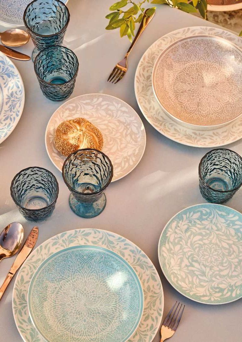 Piatti nei toni del blu pastello e del bianco e bicchieri decorati disposti su un tavolo illuminato da una luce soffusa.