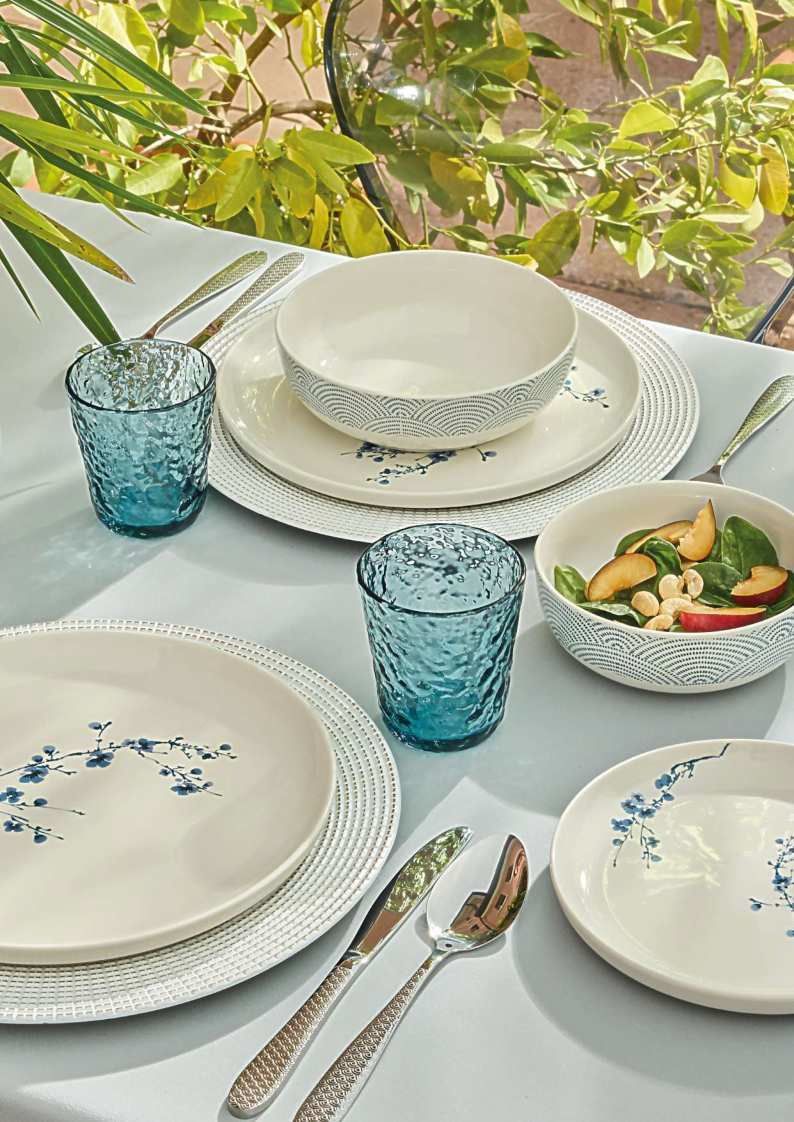 Elegante allestimento da tavola all'aperto con piatti bianchi decorati con motivi floreali, bicchieri blu e una piccola insalatiera accanto a piante verdi.