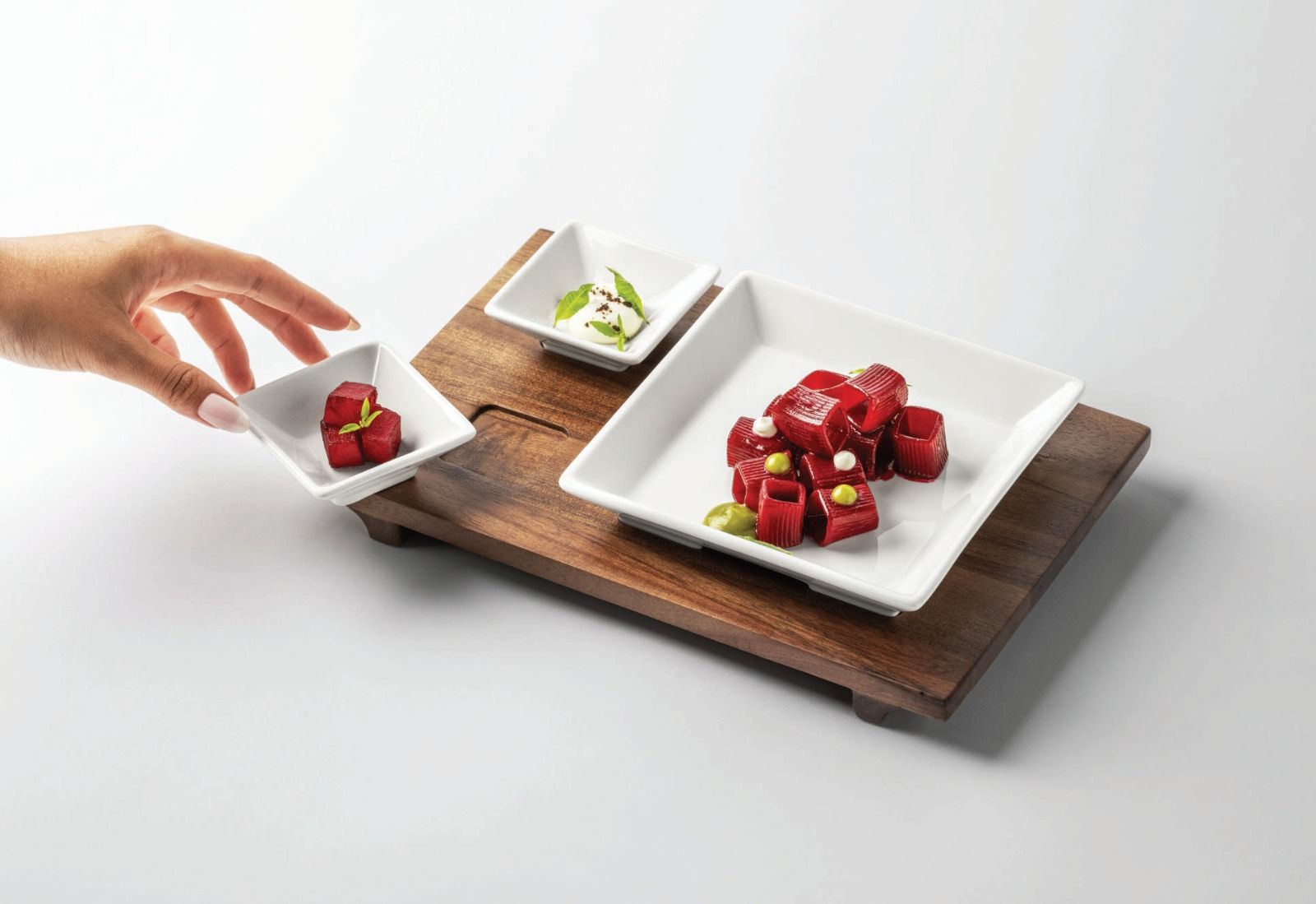 Una mano posiziona un piccolo dessert su un vassoio di legno insieme a tre piattini bianchi con frutta rossa e panna.