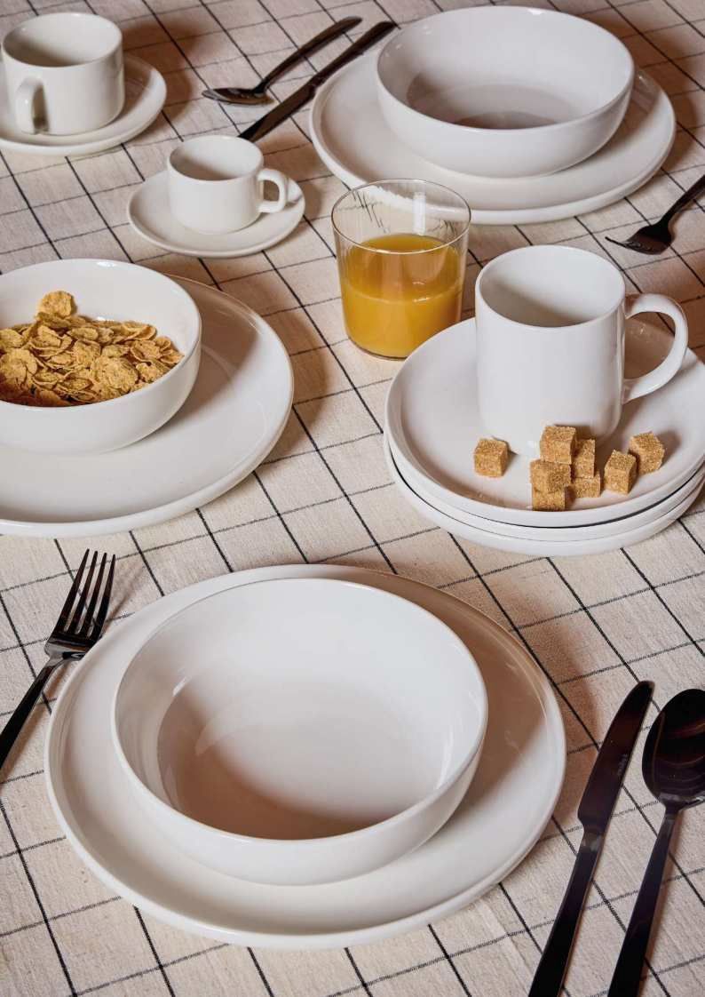 Tavolo della colazione con piatti bianchi, ciotole, tazze, cereali, succo d'arancia e posate su una tovaglia a quadri.