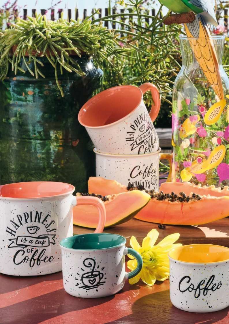 Tazze e tazzine da caffè colorate disposte su un tavolo con fiori e piante sullo sfondo.