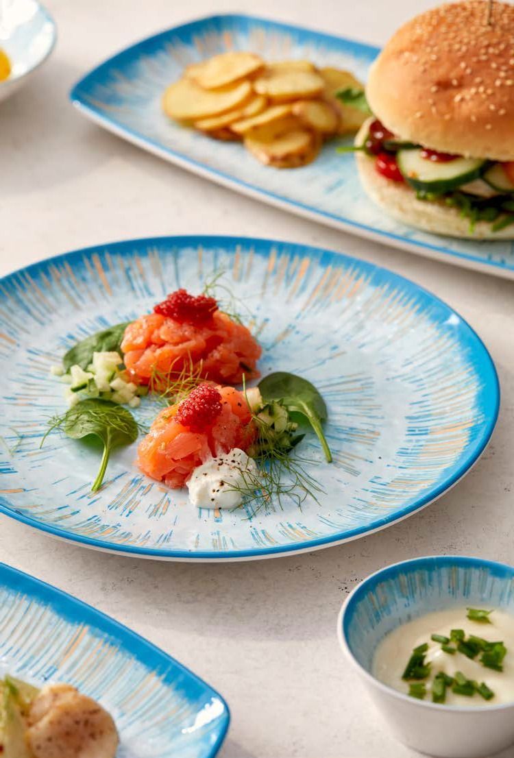 Antipasti gourmet a base di pesce serviti su piatti con bordo blu, con un hamburger e patatine fritte sullo sfondo.