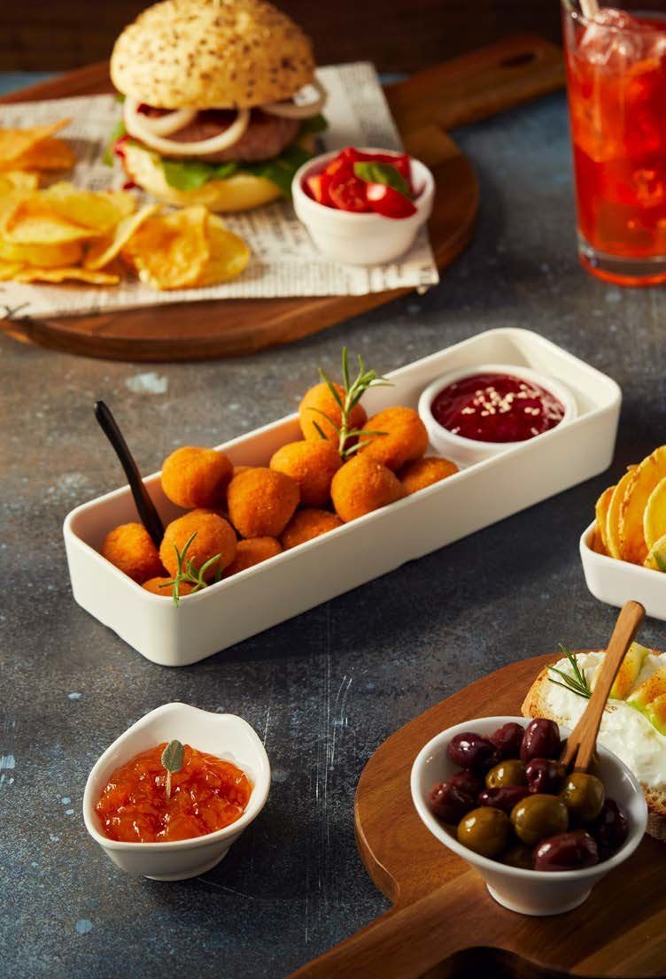 Su un tavolo scuro si trovano vari antipasti e salse, con un hamburger e una bibita ghiacciata sullo sfondo.