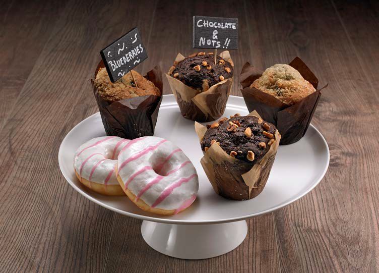 Muffin e ciambelle assortiti su un'alzatina bianca, con piccole etichette, appoggiata su un tavolo di legno.