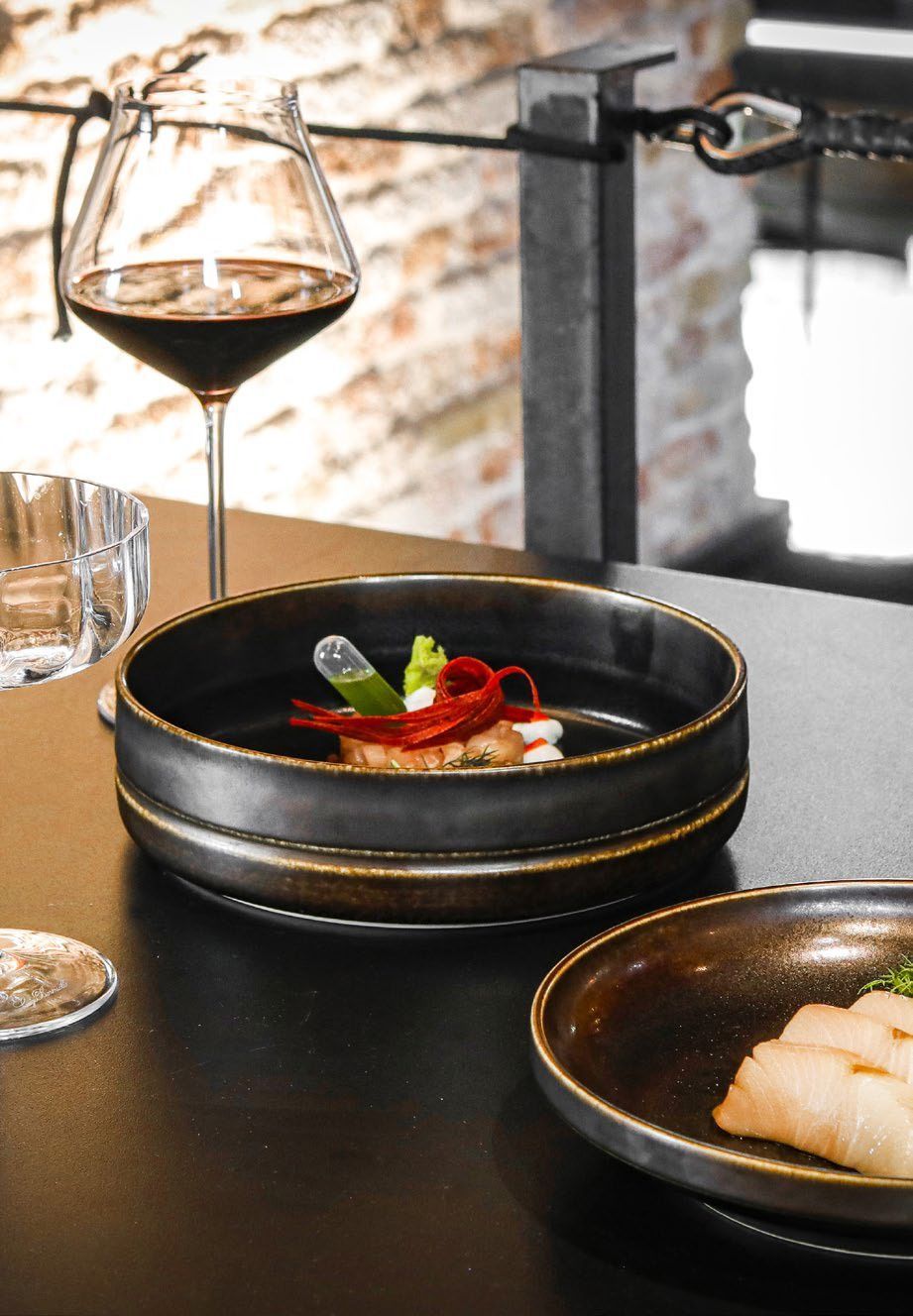 Un calice di vino accanto a piatti neri con pietanze gourmet su un tavolo in un ristorante.