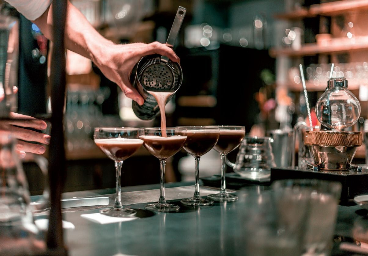 Un barista versa un cocktail scuro in una fila di bicchieri da martini in un bar.