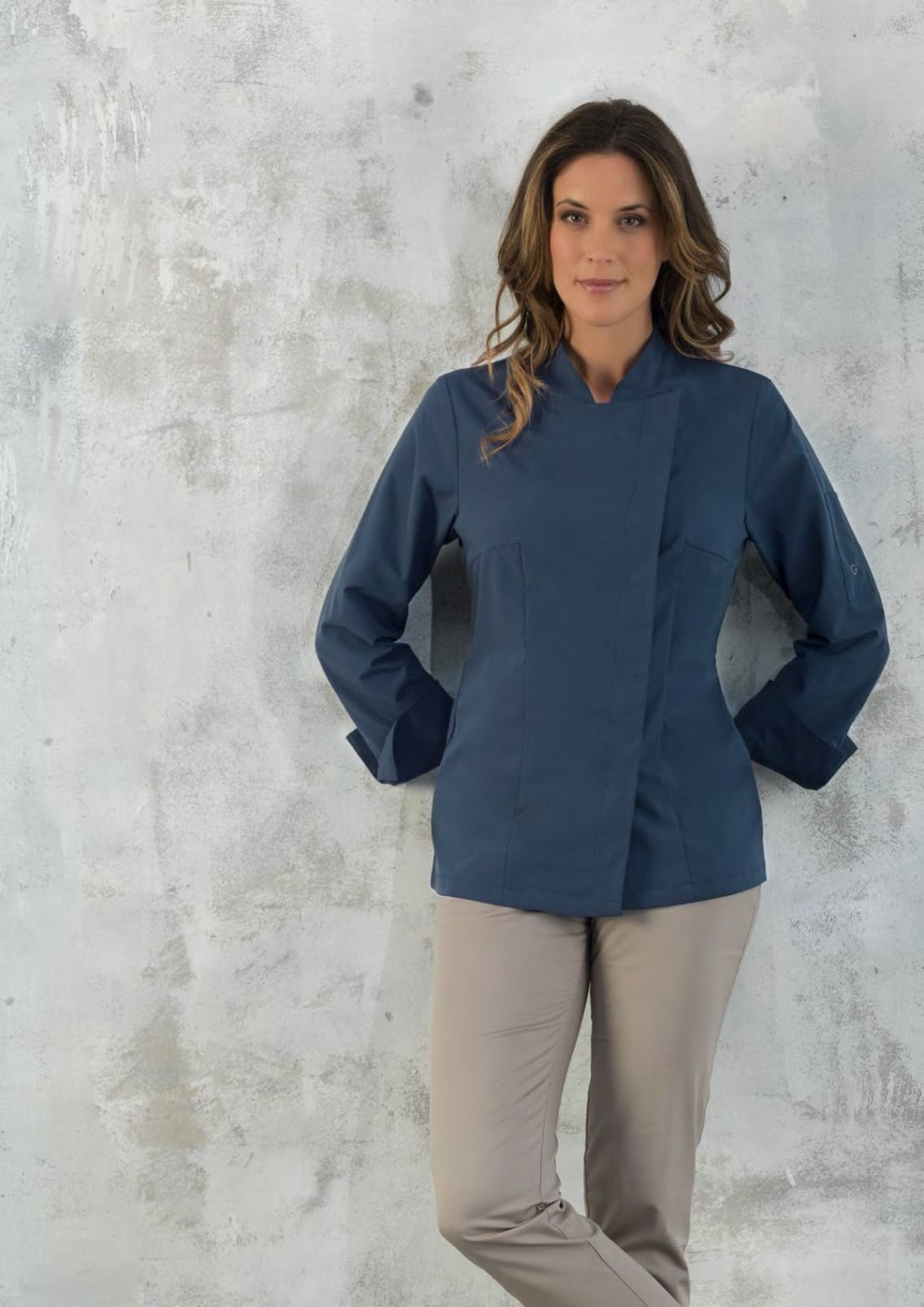 Persona con camicia blu e pantaloni beige in piedi davanti a una parete grigia con superficie ruvida.