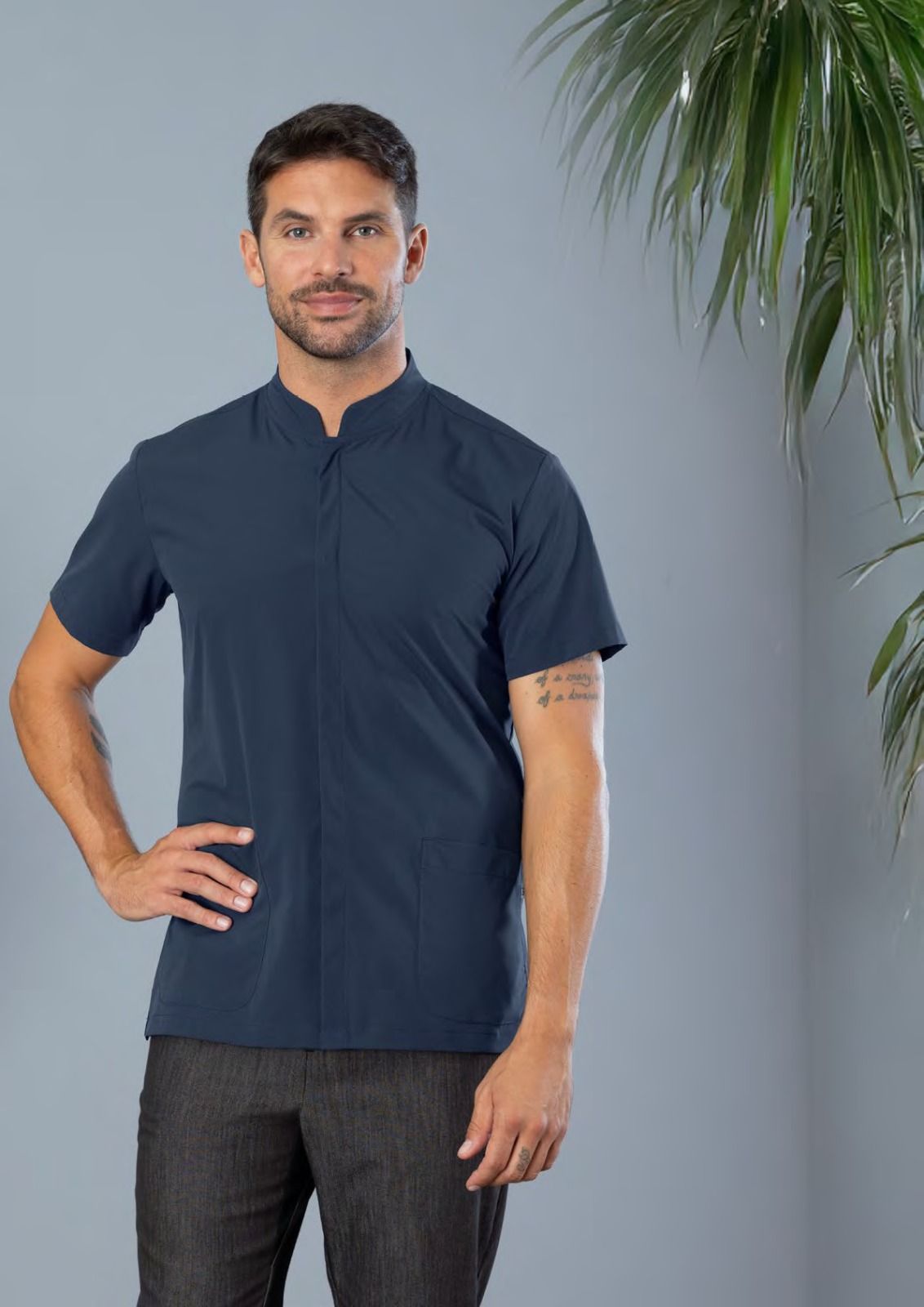 Uomo in piedi con polo blu scuro e pantaloni scuri su sfondo grigio, mano sul fianco.