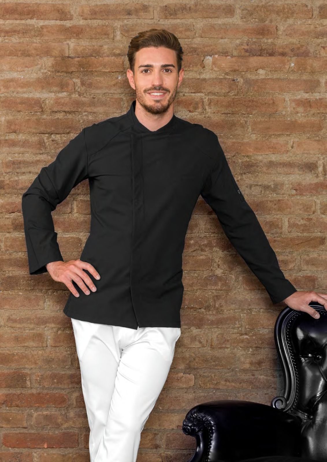 Un uomo in piedi contro un muro di mattoni, con indosso una giacca nera e pantaloni bianchi, si appoggia a una sedia nera.