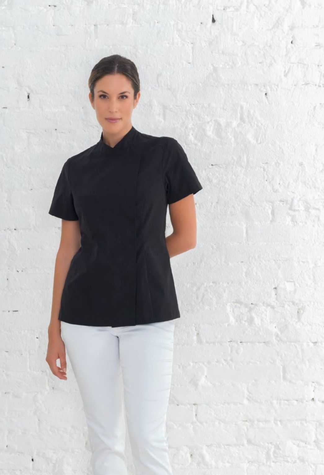 Donna con top nero e pantaloni bianchi in piedi davanti a un muro bianco.