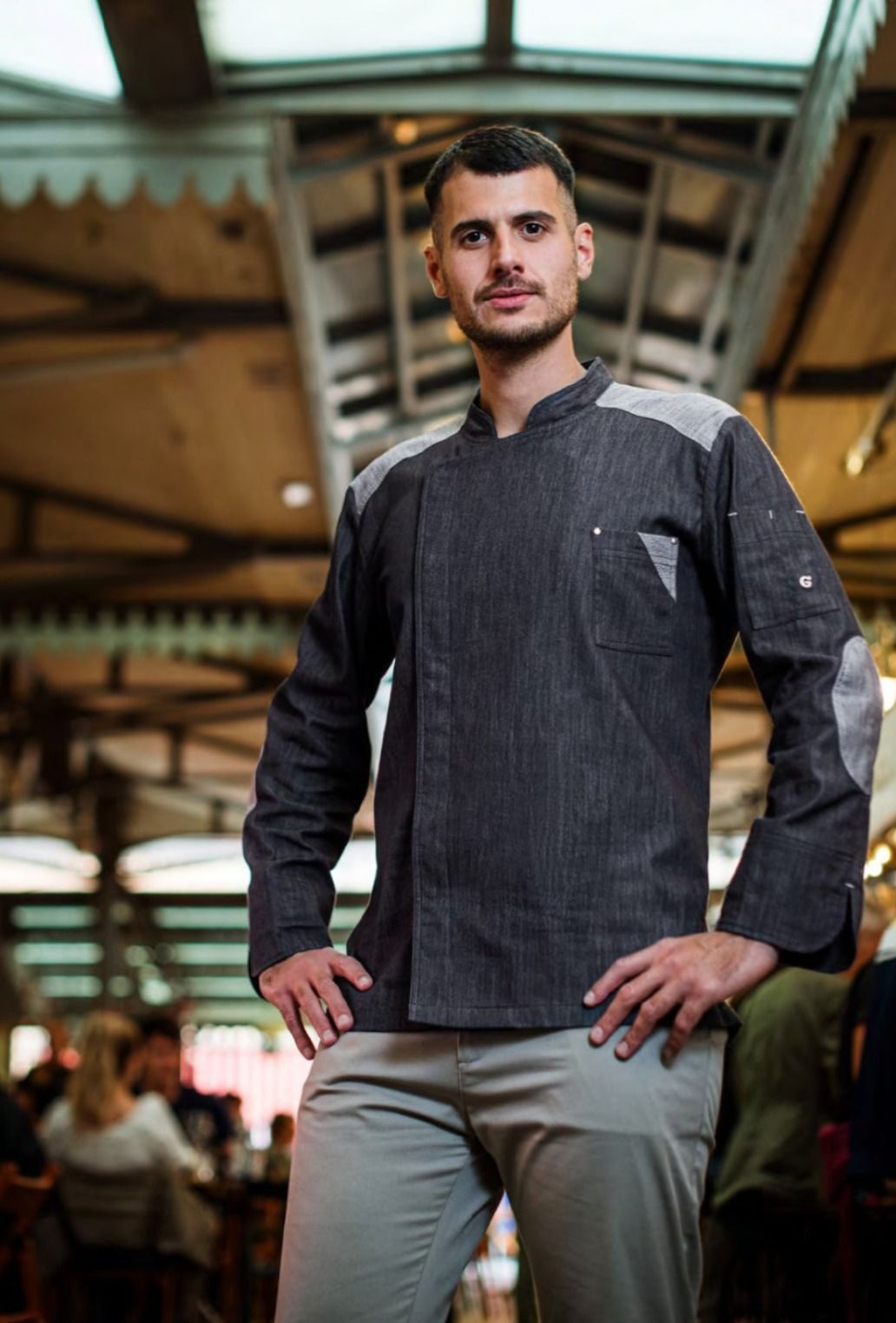 Uomo in piedi in un ambiente industriale, con indosso una camicia grigio scuro e pantaloni chiari, mani sui fianchi.