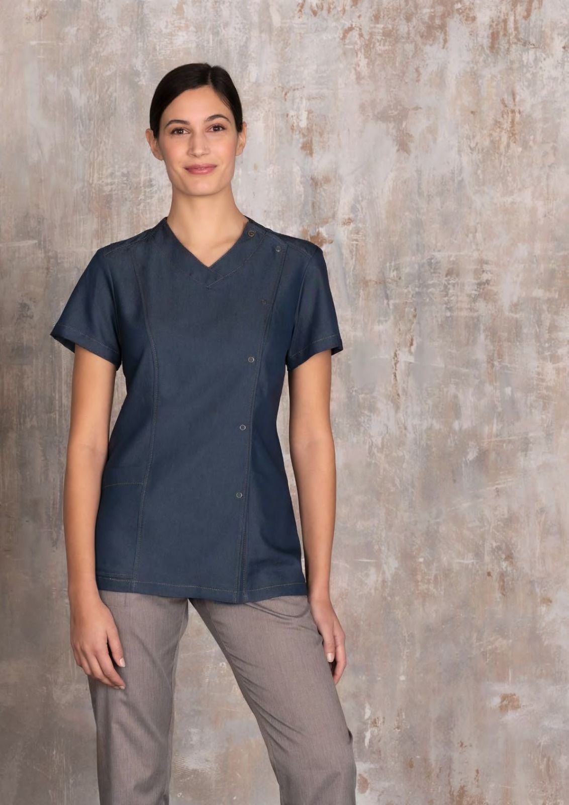 Persona con camice blu scuro e pantaloni grigi in piedi davanti a una parete beige con superficie ruvida.