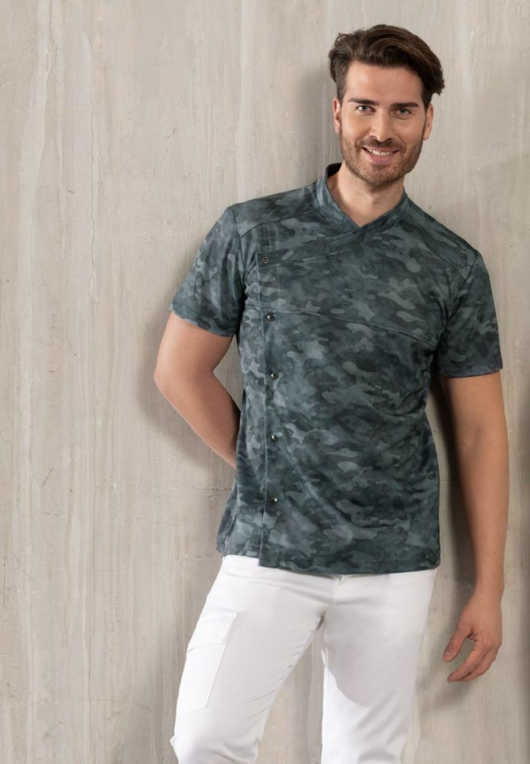 Un uomo in piedi contro un muro chiaro, con indosso una maglietta color verde acqua e pantaloni bianchi, sorride con le mani dietro la schiena.