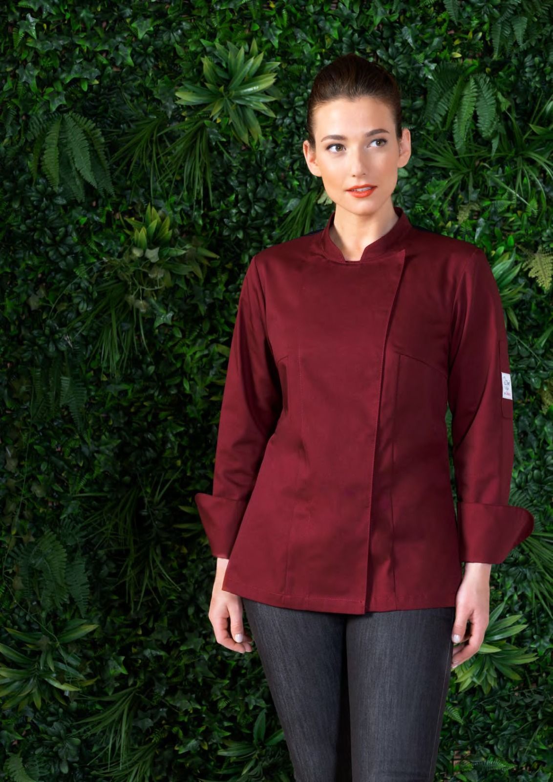 Donna con camicetta bordeaux e pantaloni scuri in piedi davanti a un muro verde ricoperto di foglie.