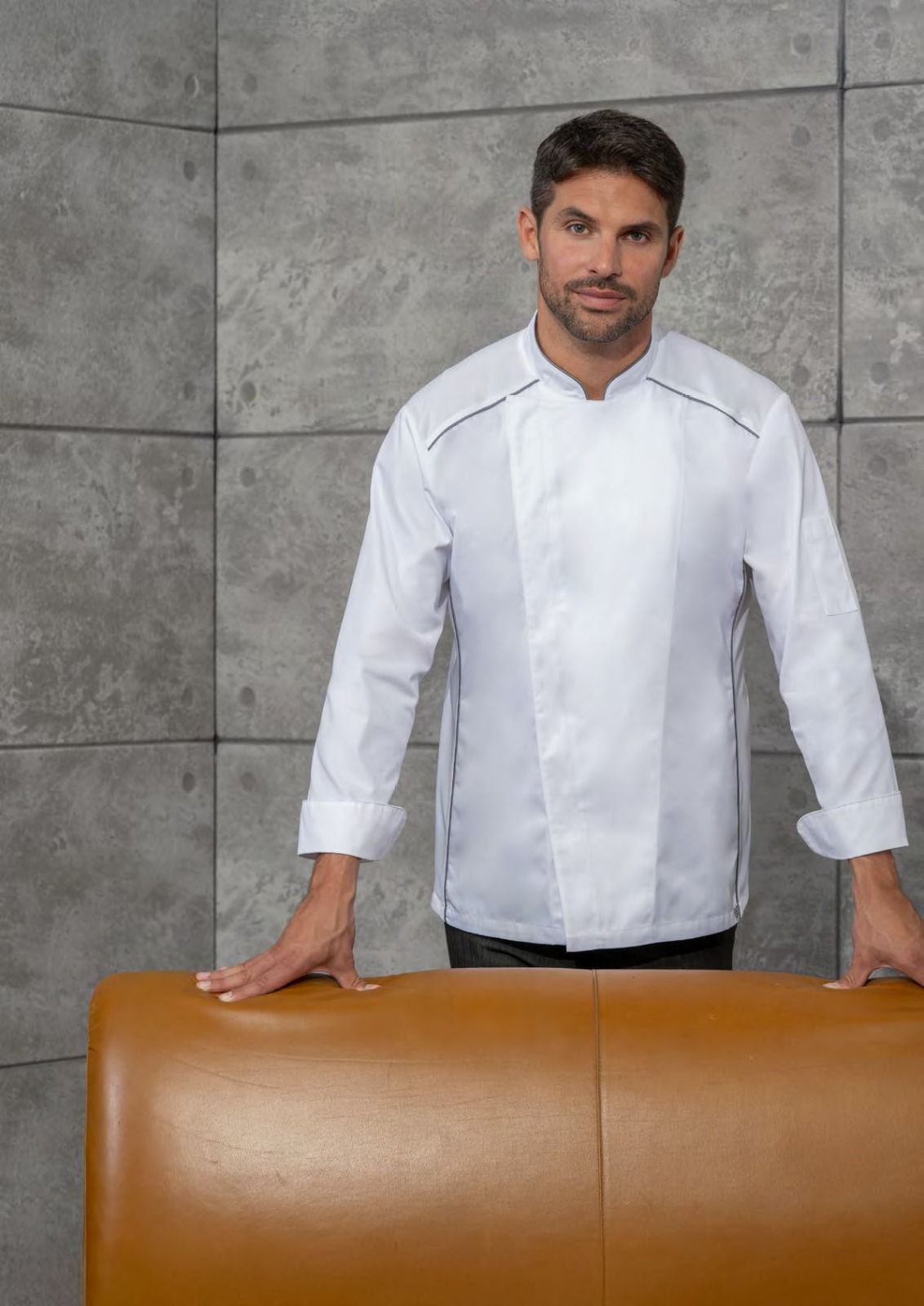 Uomo con camicia bianca in piedi dietro una poltrona di pelle color cuoio, di fronte a una parete piastrellata di grigio.