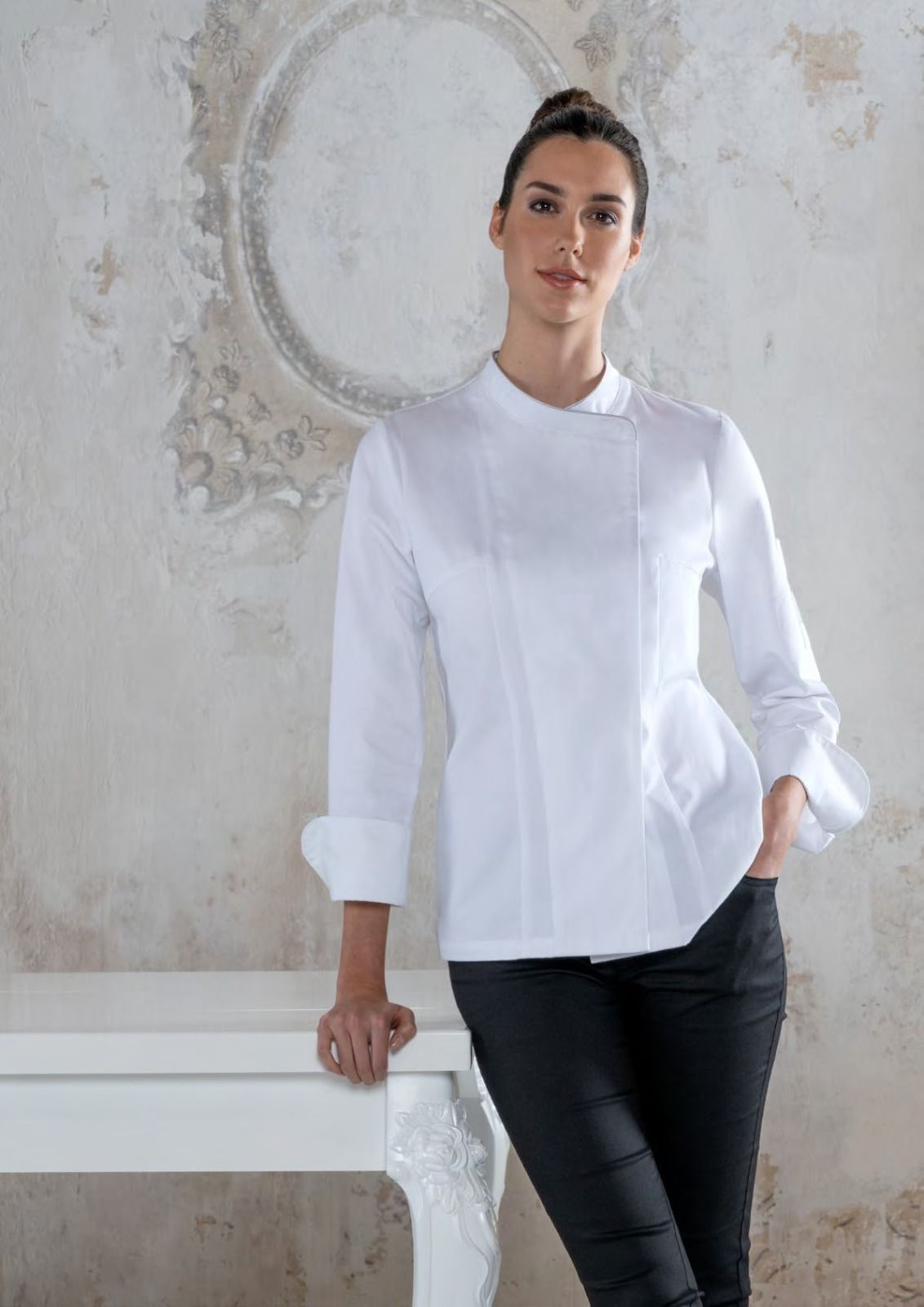 Una donna con una camicetta bianca e pantaloni neri è in piedi accanto a un tavolo bianco in una stanza luminosa e ricca di texture.