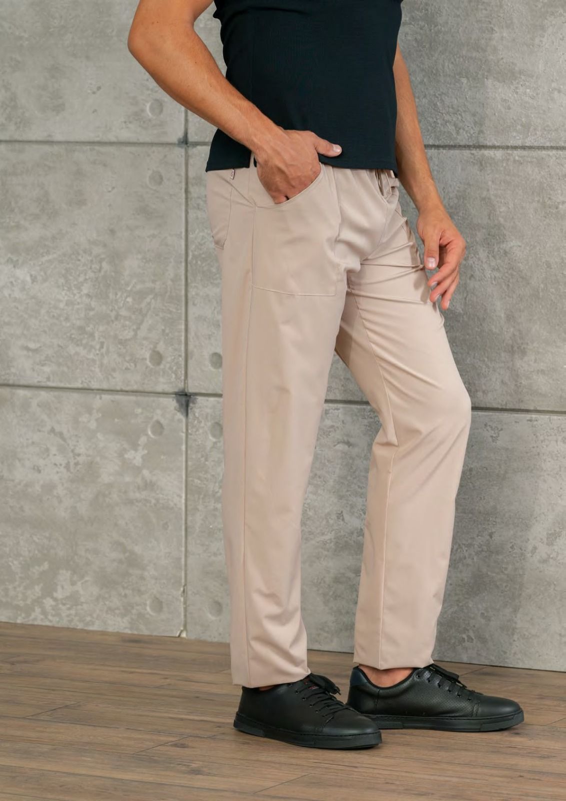 Persona in piedi con pantaloni beige, camicia nera e scarpe nere, davanti a un muro grigio.