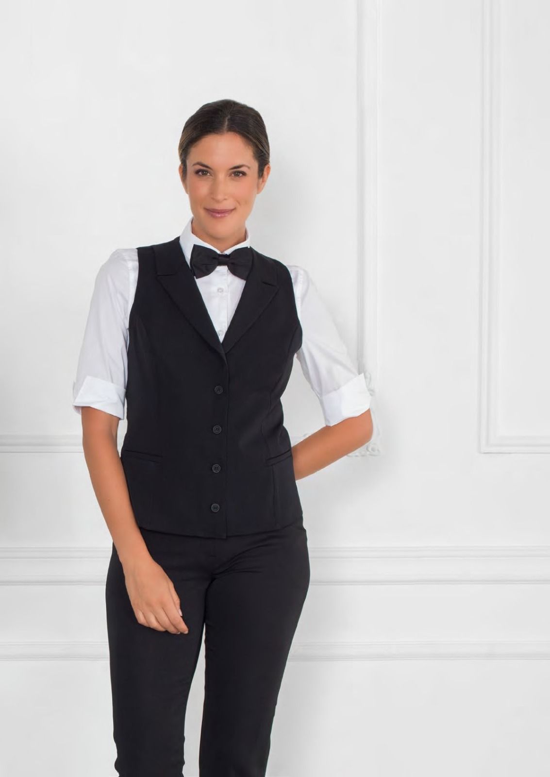 Cameriere con gilet nero, camicia bianca e papillon in piedi davanti a una parete bianca.