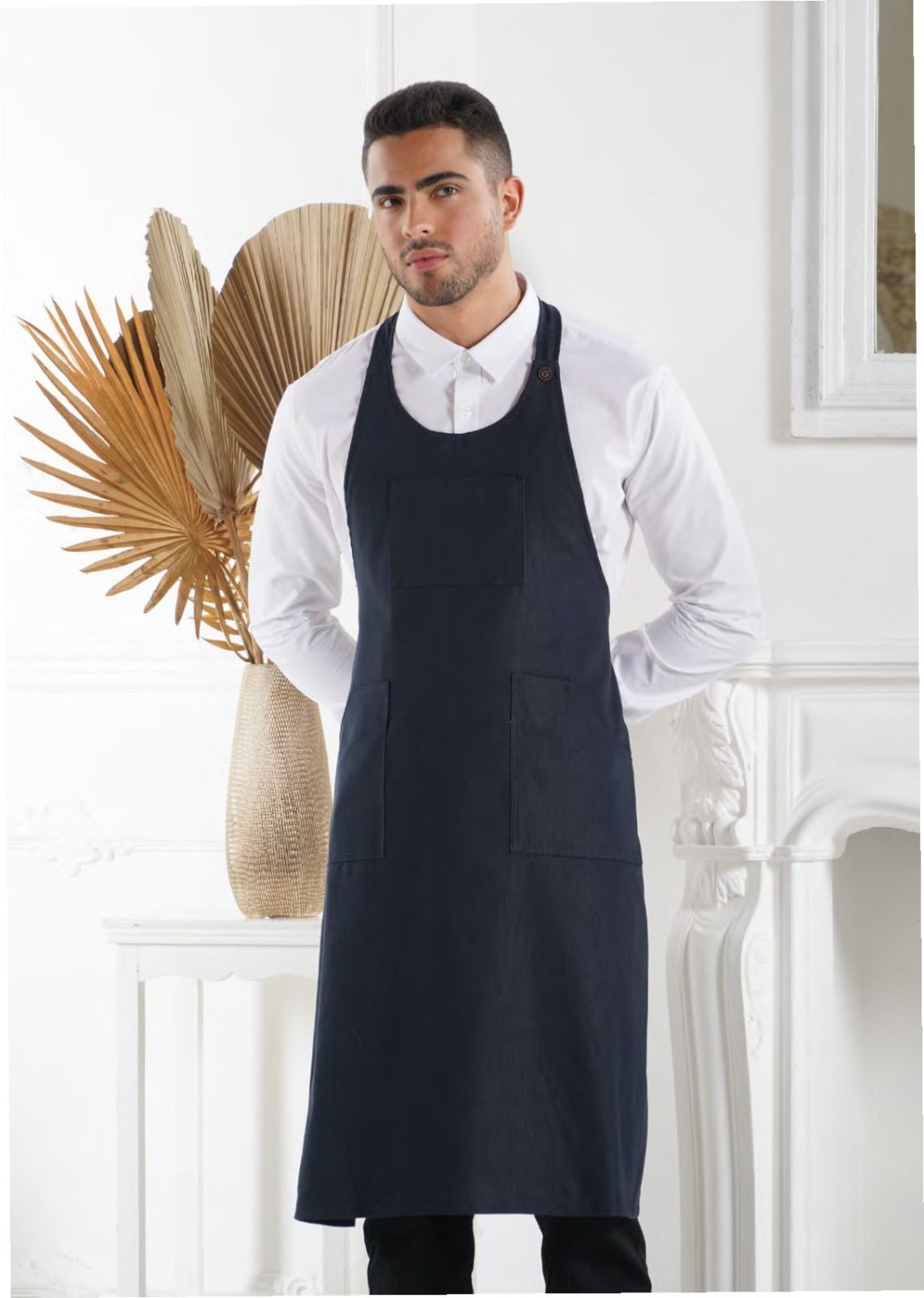 Un uomo con un grembiule blu scuro sopra una camicia bianca, in piedi in un ambiente interno accanto a un vaso decorativo e a delle foglie secche.