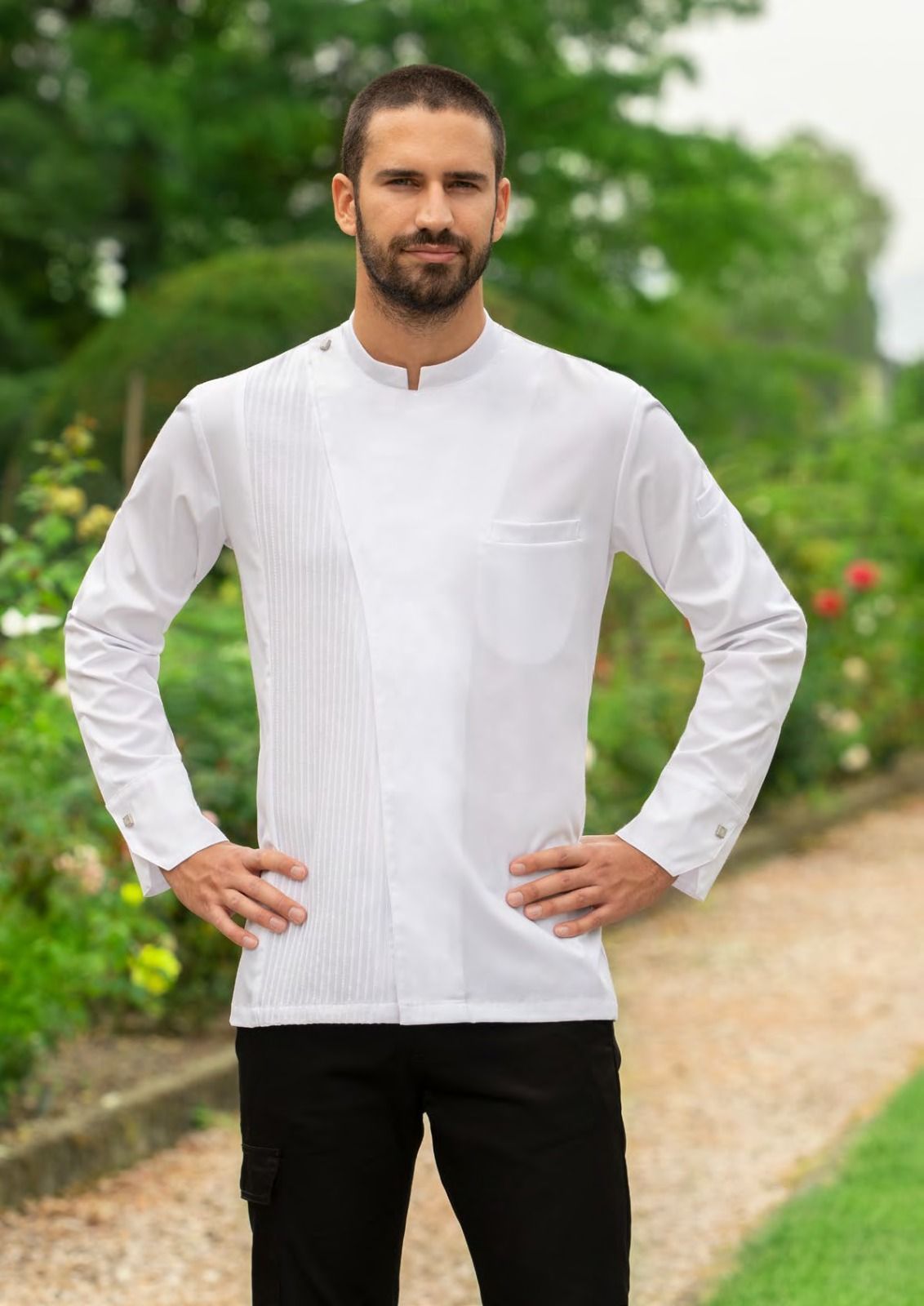 Un uomo con una camicia bianca e pantaloni neri è in piedi all'aperto, con le mani sui fianchi, lungo un sentiero in giardino.